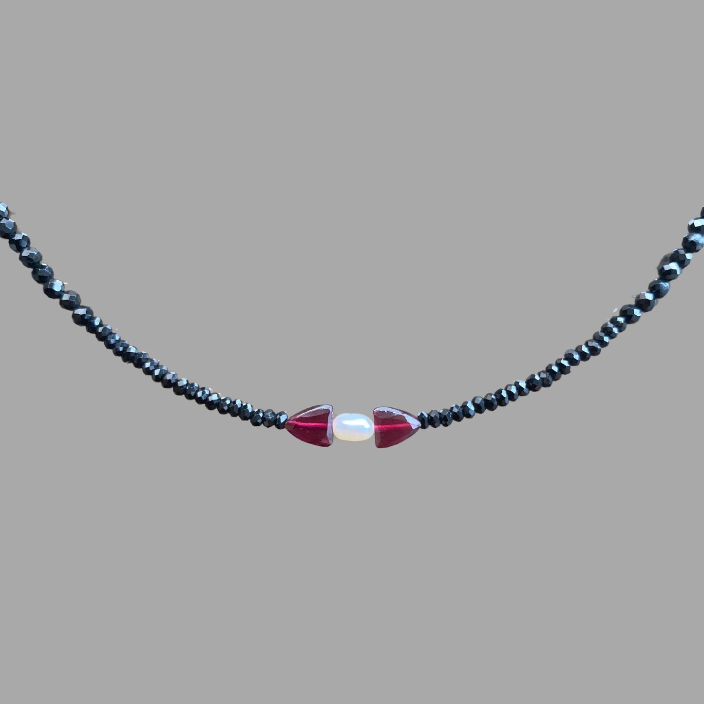 Close-up of beaded necklace, showing one rice pearl with a garnet stone on each side on a strand of black tourmaline beads.