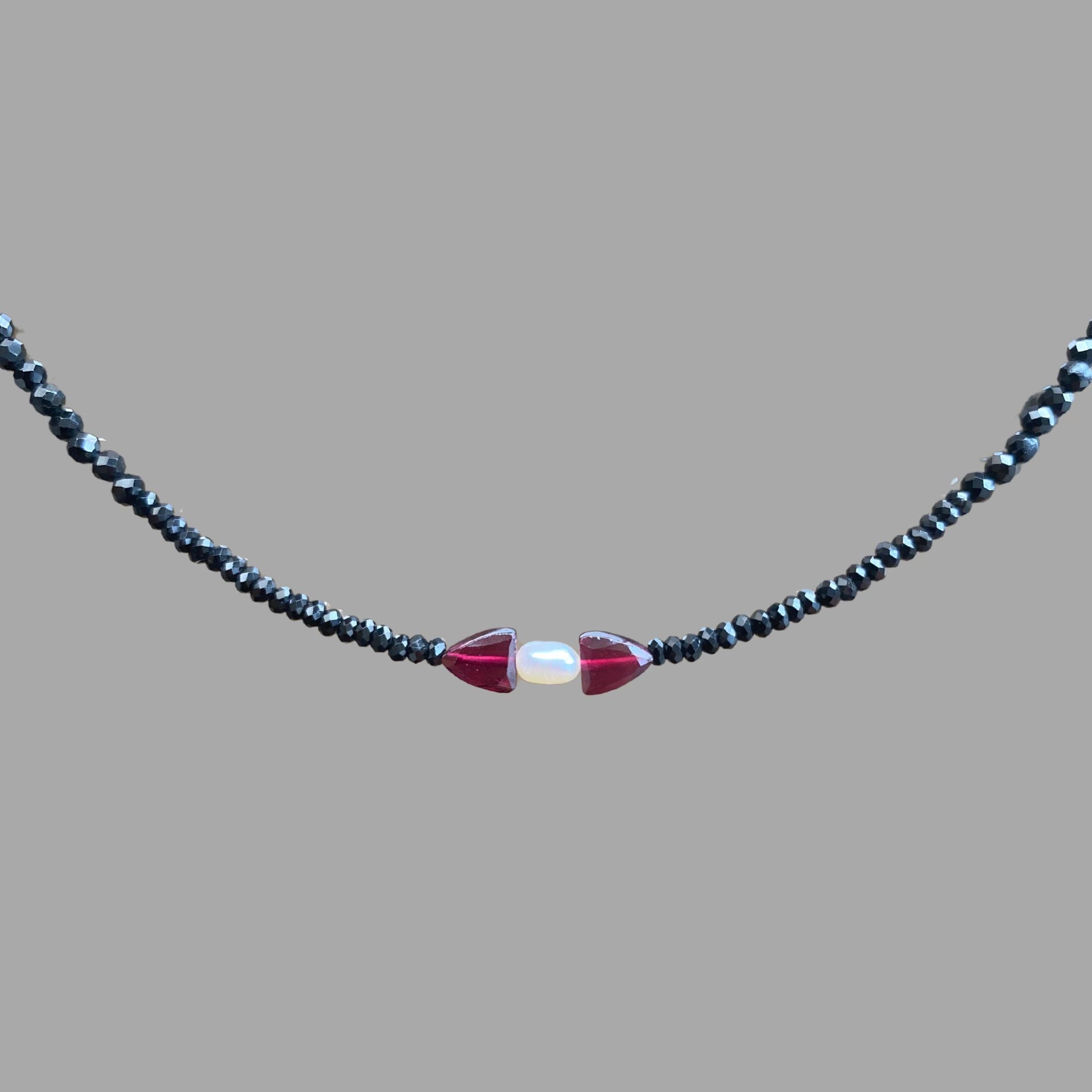 Close-up of beaded necklace, showing one rice pearl with a garnet stone on each side on a strand of black tourmaline beads.