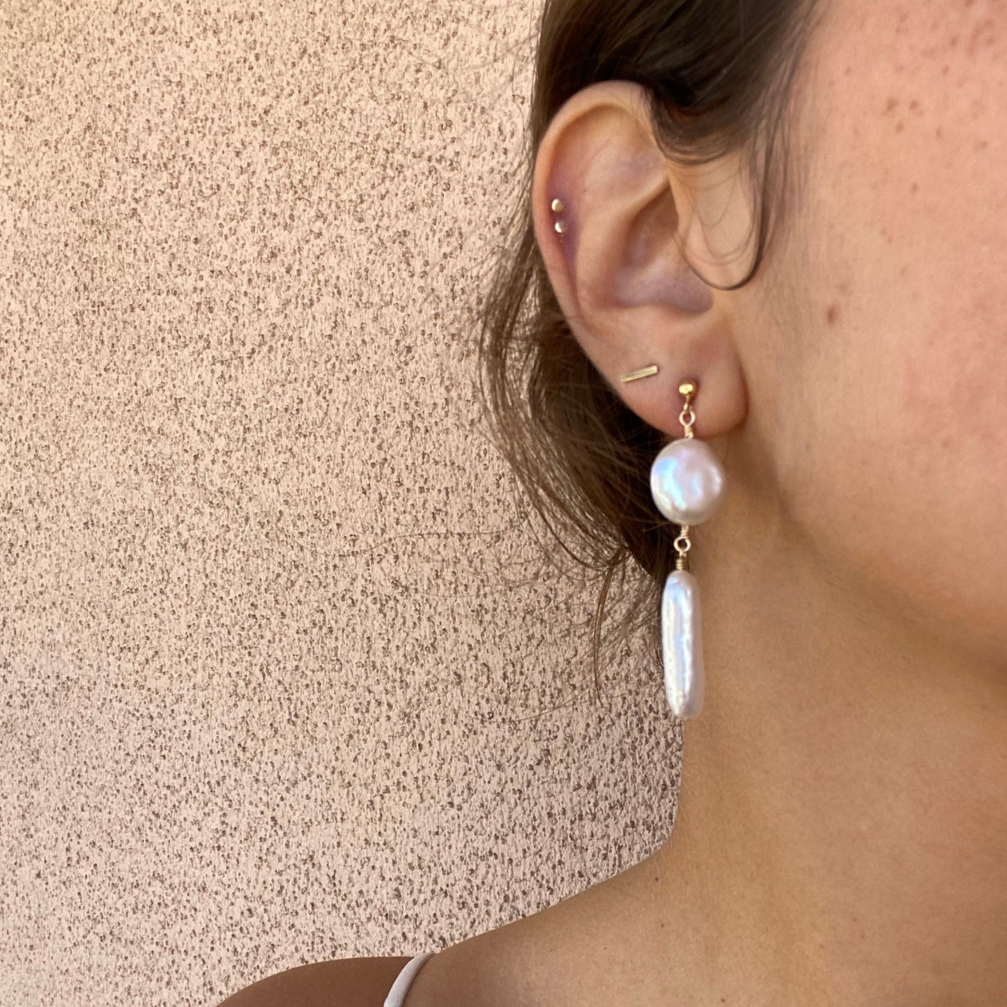 Close-up of stud earrings on a model, with only the model’s ear visible. Pearl earrings featuring a coin pearl and stick pearl hanging below the stud.