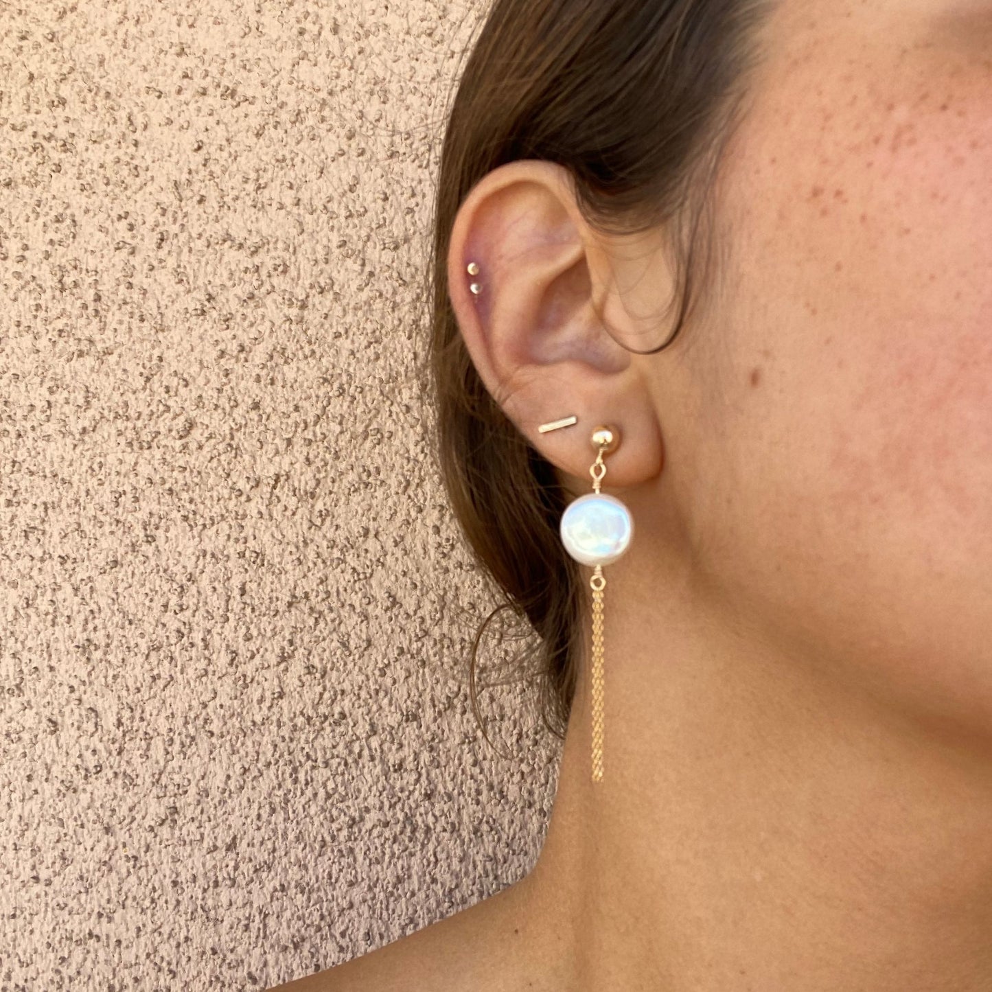 Close-up of stud earrings on a model, featuring a coin pearl with two strands of gold chain hanging below. Only the model’s ear is visible.