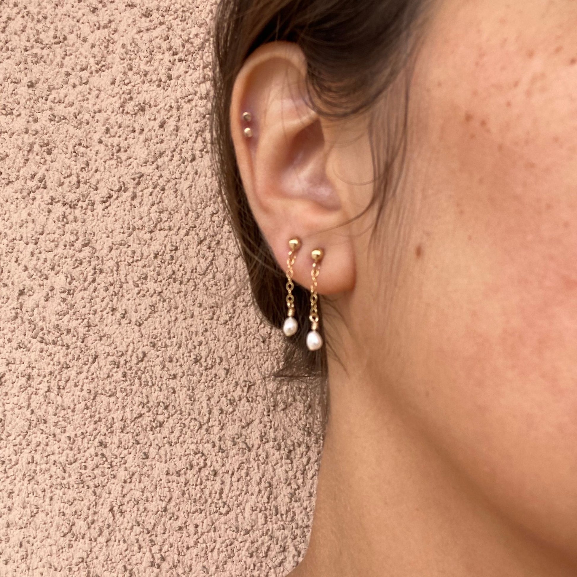 Close-up of stud earrings on a model, featuring a short gold chain segment with a small rice pearl at the bottom. Only the model’s ear is visible.