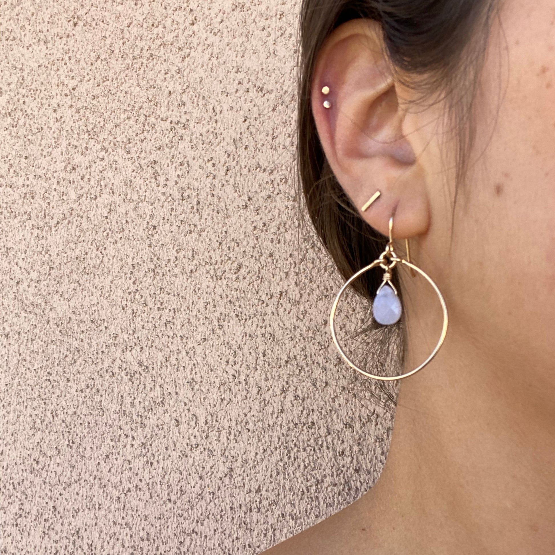 Close-up of gold hoop earrings on a model, featuring a blue chalcedony gem hanging from the top center of the hoop. Only the model’s ear is visible.