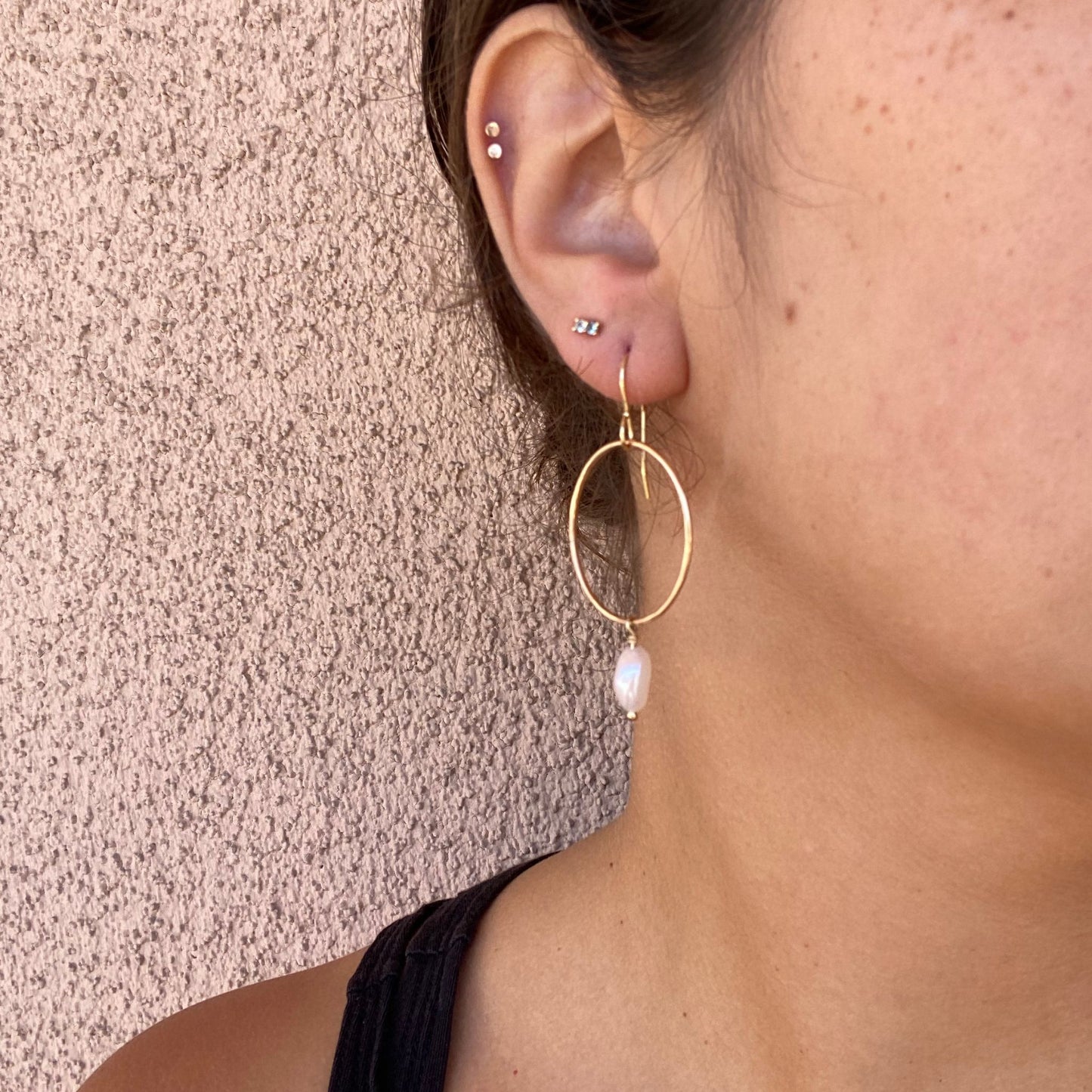 Close-up of gold hoop earrings on a model, featuring a freshwater pearl hanging from the bottom of the hoop. Only the model’s ear is visible.