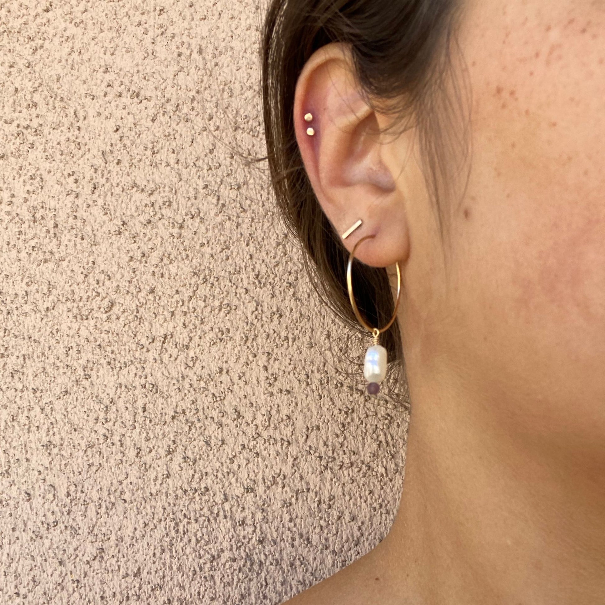 Close-up of gold hoop earring on a model, with a freshwater pearl and a purple sapphire bead hanging from the hoop. Only the model’s ear is visible.