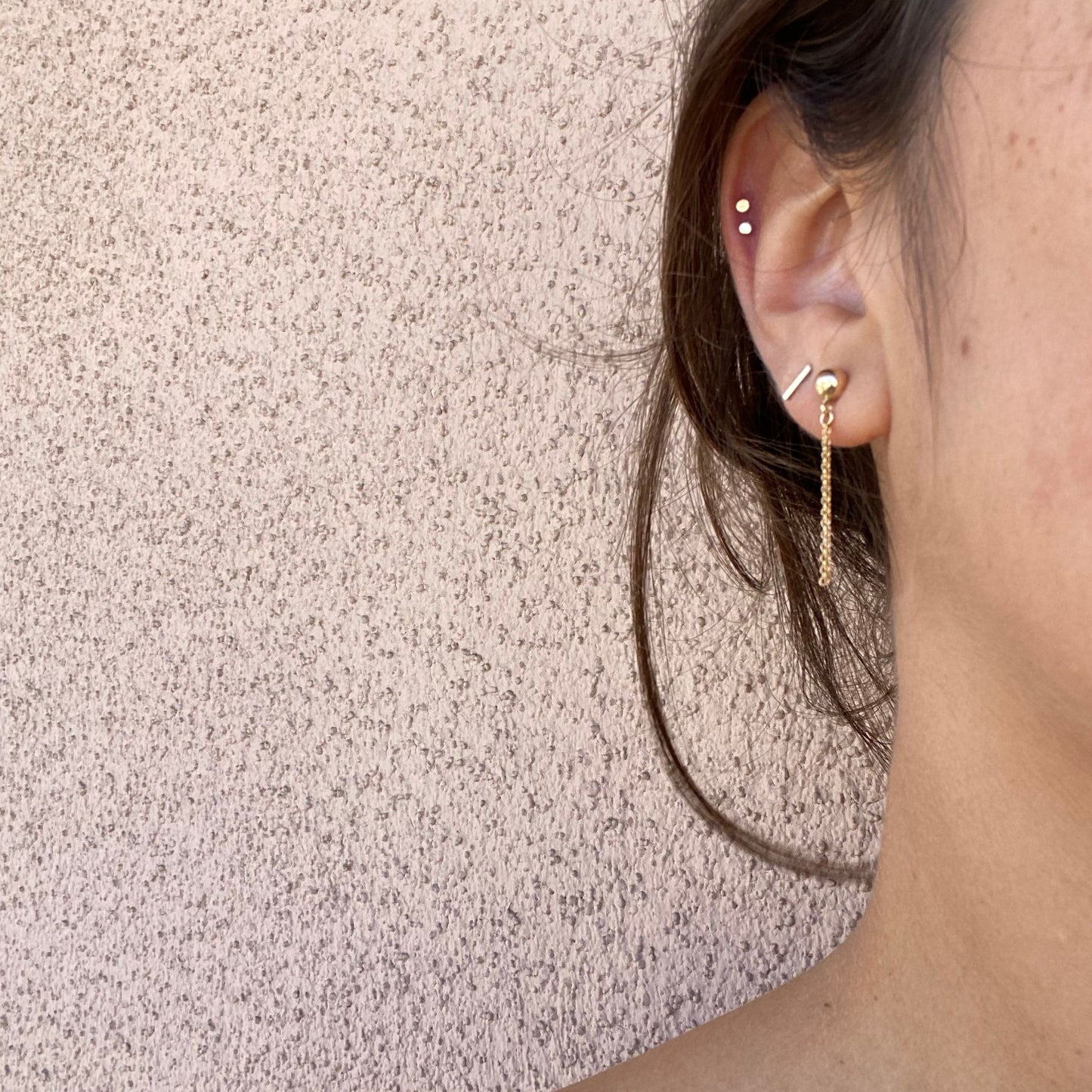 Close-up of stud earrings on a model, a gold ball stud with two strands of gold chain hanging below the ball. Only the model’s ear is visible.