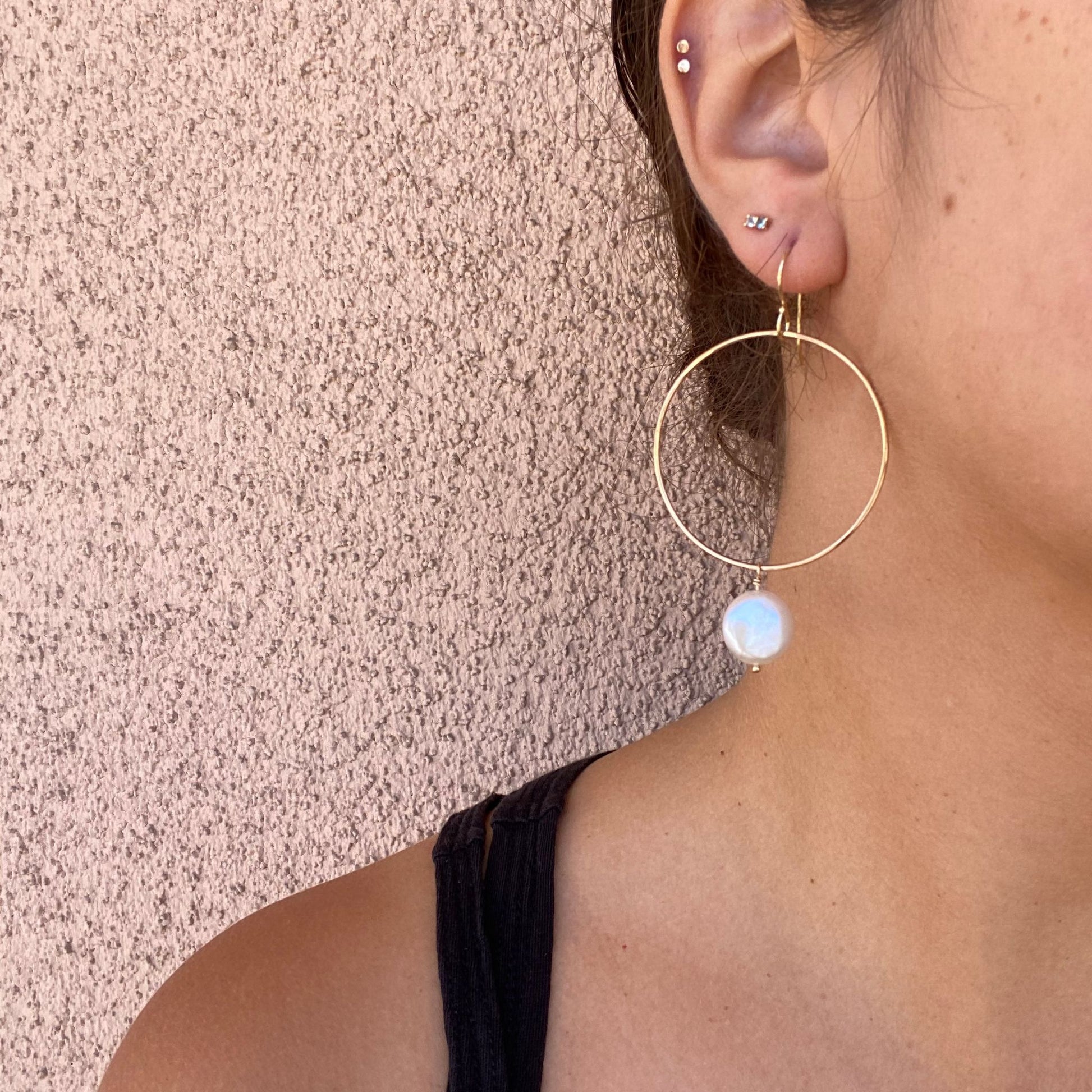 Close-up of gold hoop earring on a model, featuring a round coin pearl hanging from the bottom of the hoop. Only the model’s ear is visible.