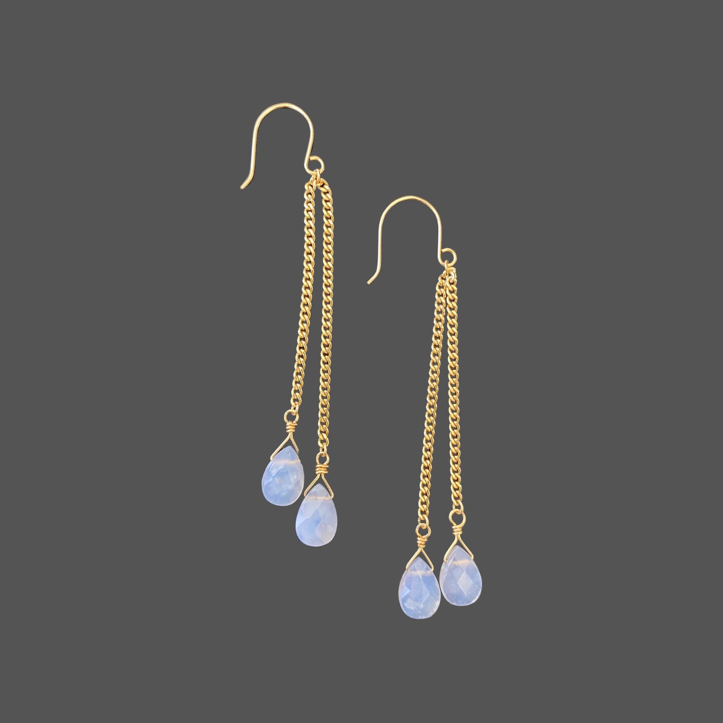 Side view of long earrings on French earwires, with asymmetrical gold chains ending in blue chalcedony gemstones. Shown alone on plain background.