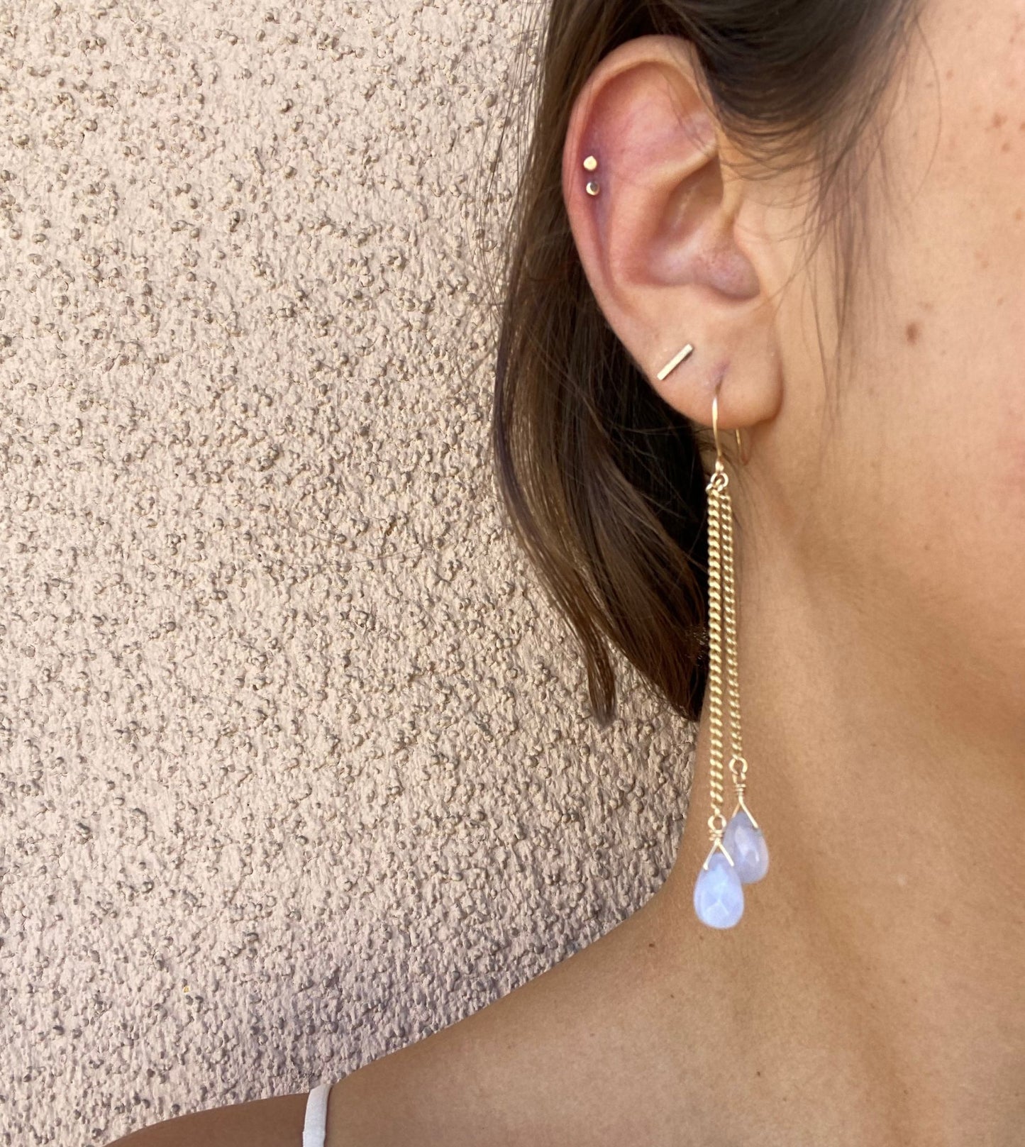 Long earrings shown on model, with asymmetrical gold chains ending in blue chalcedony gemstones. Only the model’s ear is visible.